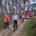 W tej scenie uczestnicy marszu nordic walking poruszają się po leśnej ścieżce. Za nimi widać wóz strażacki, który czuwa nad bezpieczeństwem uczestników.