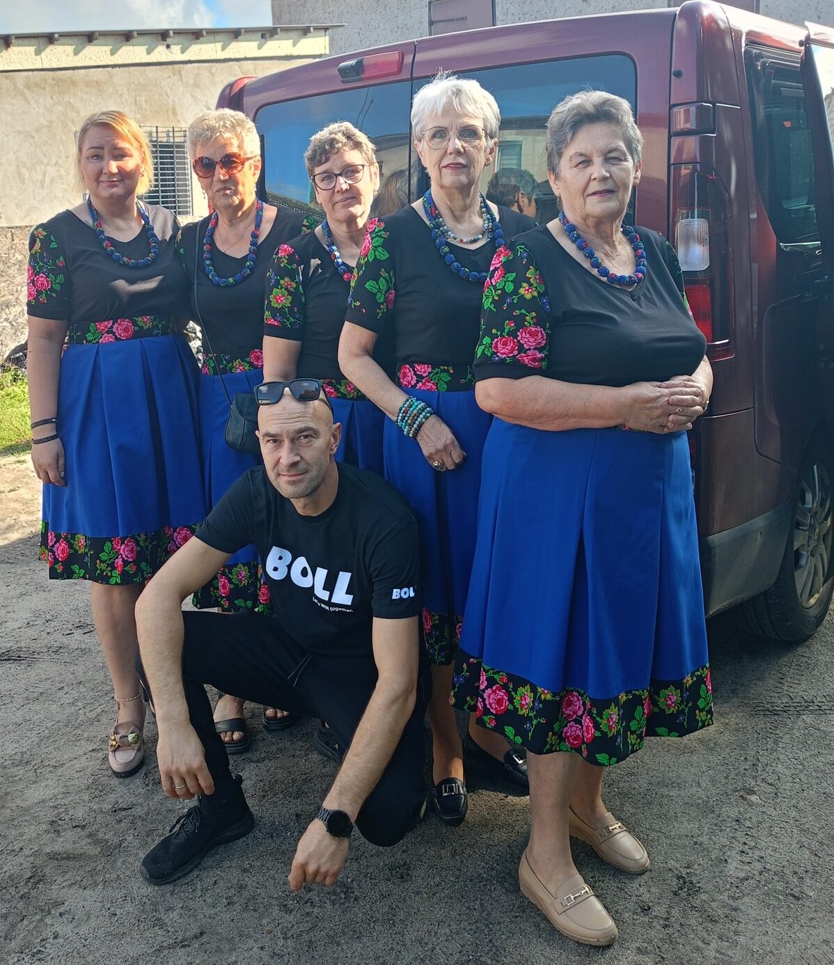 Na zdjęciu widzimy grupę sześciu osób – pięć kobiet i mężczyznę, którzy stoją razem na tle samochodu. Kobiety są ubrane w tradycyjne stroje z kwiatowym wzorem na dole spódnic, z koralami na szyjach, co nadaje im regionalny charakter.