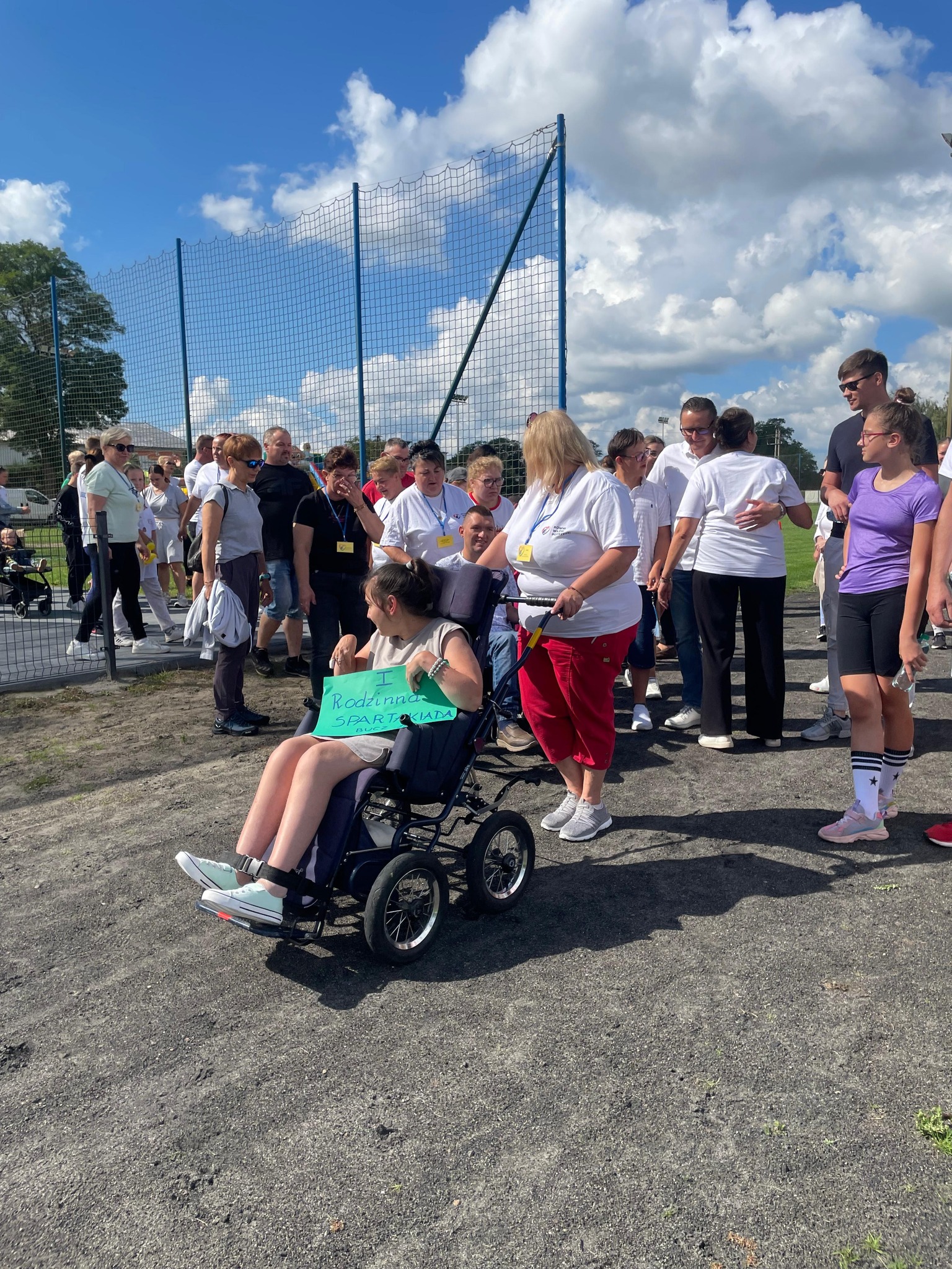 Na zdjęciu widzimy grupę uczestników I Rodzinnej Spartakiady, którzy są w trakcie przygotowań do konkurencji. Wśród nich znajduje się osoba na wózku inwalidzkim, której towarzyszy opiekun.