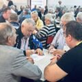 Na zdjęciu widzimy salę wypełnioną osobami, które siedzą przy stołach i grają w karty. W centrum kadru znajduje się stół, przy którym czterech mężczyzn skupia się na grze.