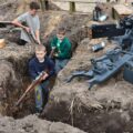 Na fotografii dzieci biorą udział w rekonstrukcji warunków frontowych – poruszają się po wykopanych okopach, trzymając repliki broni z epoki. Obok znajduje się zrekonstruowany ciężki karabin maszynowy.