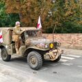 Na fotografii wojskowy pojazd sanitarny z czasów wojny porusza się ulicą podczas przejazdu historycznego. Rekonstruktorzy w mundurach sanitariuszy oraz flagi z czerwonym krzyżem podkreślają charakter pojazdu.