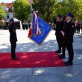 Na fotografii uroczysty moment oddania honorów sztandarowi. Po lewej stronie, na czerwonym dywanie, funkcjonariusz Państwowej Straży Pożarnej wykonuje salut. Na wprost niego stoi poczet sztandarowy trzymający sztandar Komendy Powiatowej PSP w Wolsztynie. W tle widoczna jest kolumna strażaków w umundurowaniu galowym.