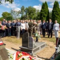Zdjęcie przedstawia uroczystość patriotyczną na cmentarzu. Na pierwszym planie stoi żołnierka w mundurze, patrząca w stronę zgromadzonych uczestników. Centralnie widoczny jest inny żołnierz, który odbiera wieniec od jednej z delegacji. W tle znajduje się liczna grupa osób – kobiety i mężczyźni w eleganckich strojach, przedstawiciele władz i urzędów.