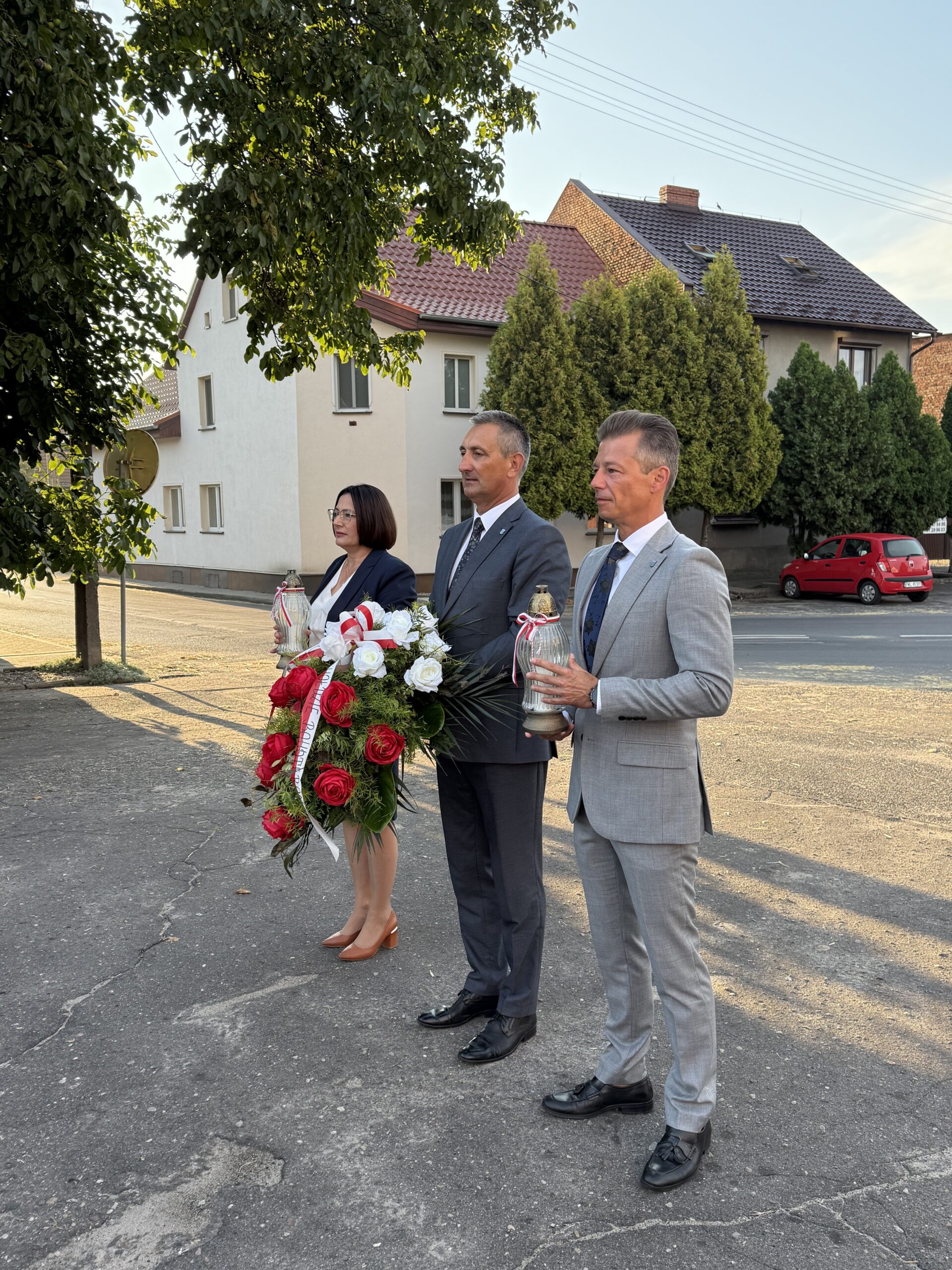 Na zdjęciu wójt, zastępca oraz skarbnik. Wszyscy stoją pod pomnikiem. Wójt trzyma wiązankę, a zastępca i skarbnik znicze.