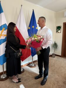 Na fotografii uchwycono moment wręczania bukietu kwiatów pani Edycie przez wójta. W tle flagi Polski, Unii Europejskiej oraz flaga z herbem gminy.