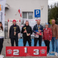 Na fotografii trójka mężczyzn pozuje z medalami i pucharami. Na fotografii także wójt i dwie inne osoby. Przed nimi znajduje się podium sportowe.