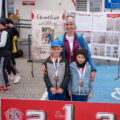 Na zdjęciu kobieta z dwoma chłopcami. Wszyscy mają założone medale. Przed nimi znajduje się podium sportowe.