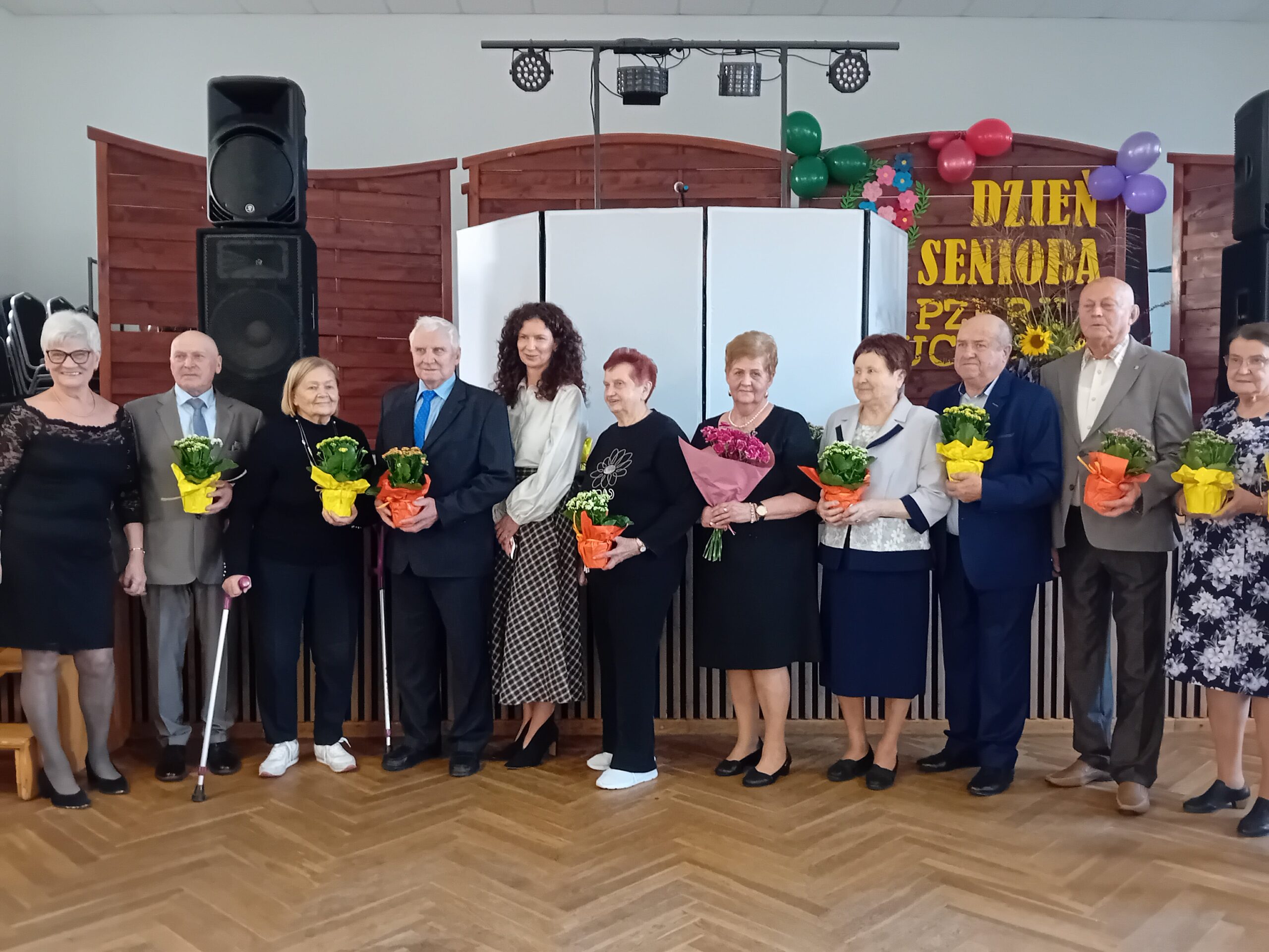 Grupowe zdjęcie osób obecnych na uroczystości. Seniorzy trzymają kwiatki w doniczkach.