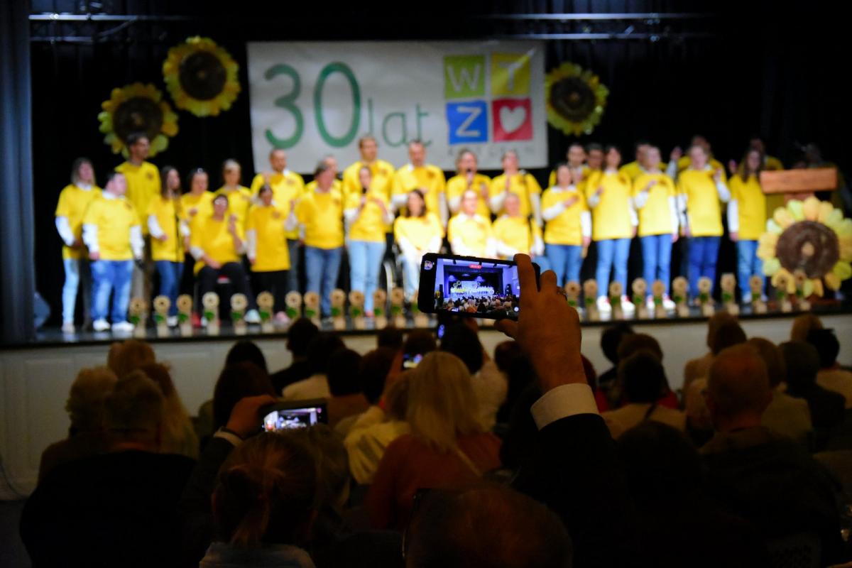 Zdjęcie zostało wykonane z tylnej części sali. Widać na nim publiczność siedzącą na widowni oraz uczestników warsztatów występujących na scenie.