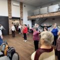 Grupa seniorów wraz z fizjoterapeutką wykonuje ćwiczenia.