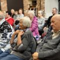 Zebrani seniorzy siedzą na krzesłach i zainteresowaniem słuchają wykładu.