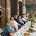 Na fotografii seniorzy siedzą przy stołach. Na stołach znajdują się ciasta i owoce, a także wazony ze słonecznikami.