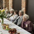 Na fotografii seniorzy siedzą przy stołach. Na stołach znajdują się ciasta i owoce, a także wazony ze słonecznikami.