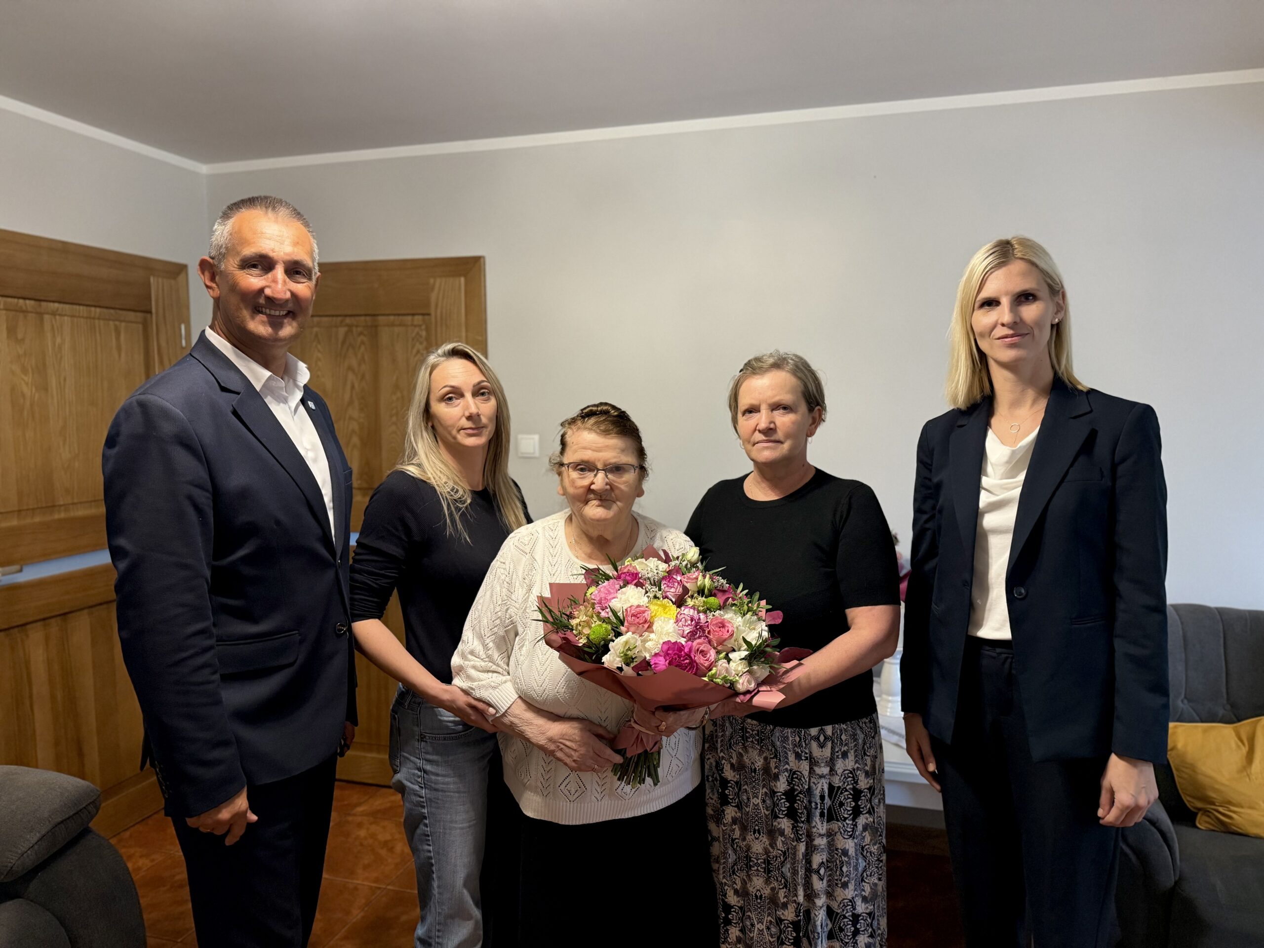 Grupowe zdjęcie, na którym znajdują się wójt, zastępca kierownika USC oraz jubilatka ze swoją rodziną. Pani Jadwiga pozuje z bukietem kwiatów.