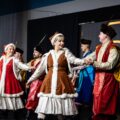 Na zdjęciu widoczni są mężczyźni i kobiety ubrani w ludowe stroje, tańczący na scenie.