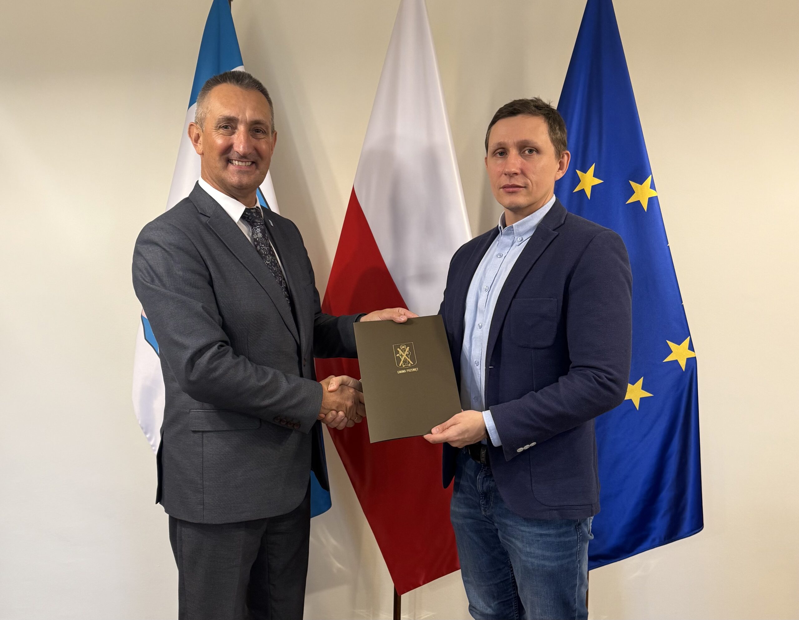 Na zdjęciu wójt oraz pan Marek Smolarek podają sobie dłoń oraz trzymają teczkę, w której znajduje się podpisana umowa. W tle flagi: Polski, Unii Europejskiej oraz flaga z herbem gminy Przemęt.