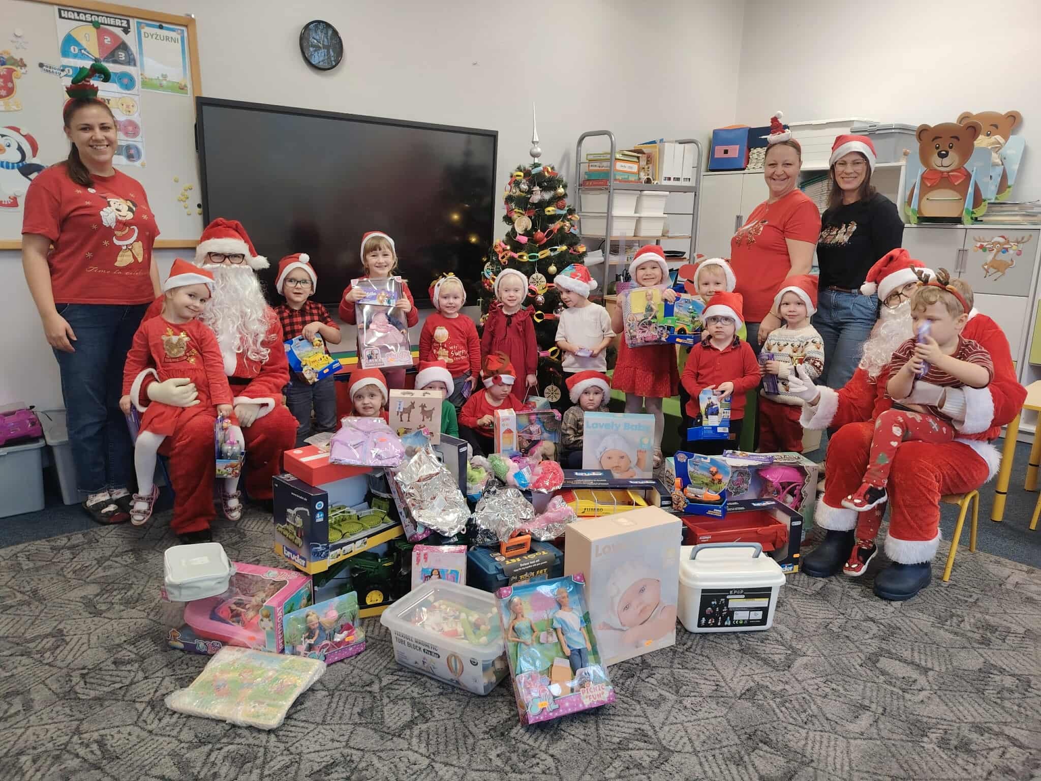 Na zdjęciu widoczne są przedszkolaki, które pozują z prezentami. Na fotografii znajdują się także dyrektorka szkoły wraz z nauczycielkami oraz osoba przebrana za Świętego Mikołaja.