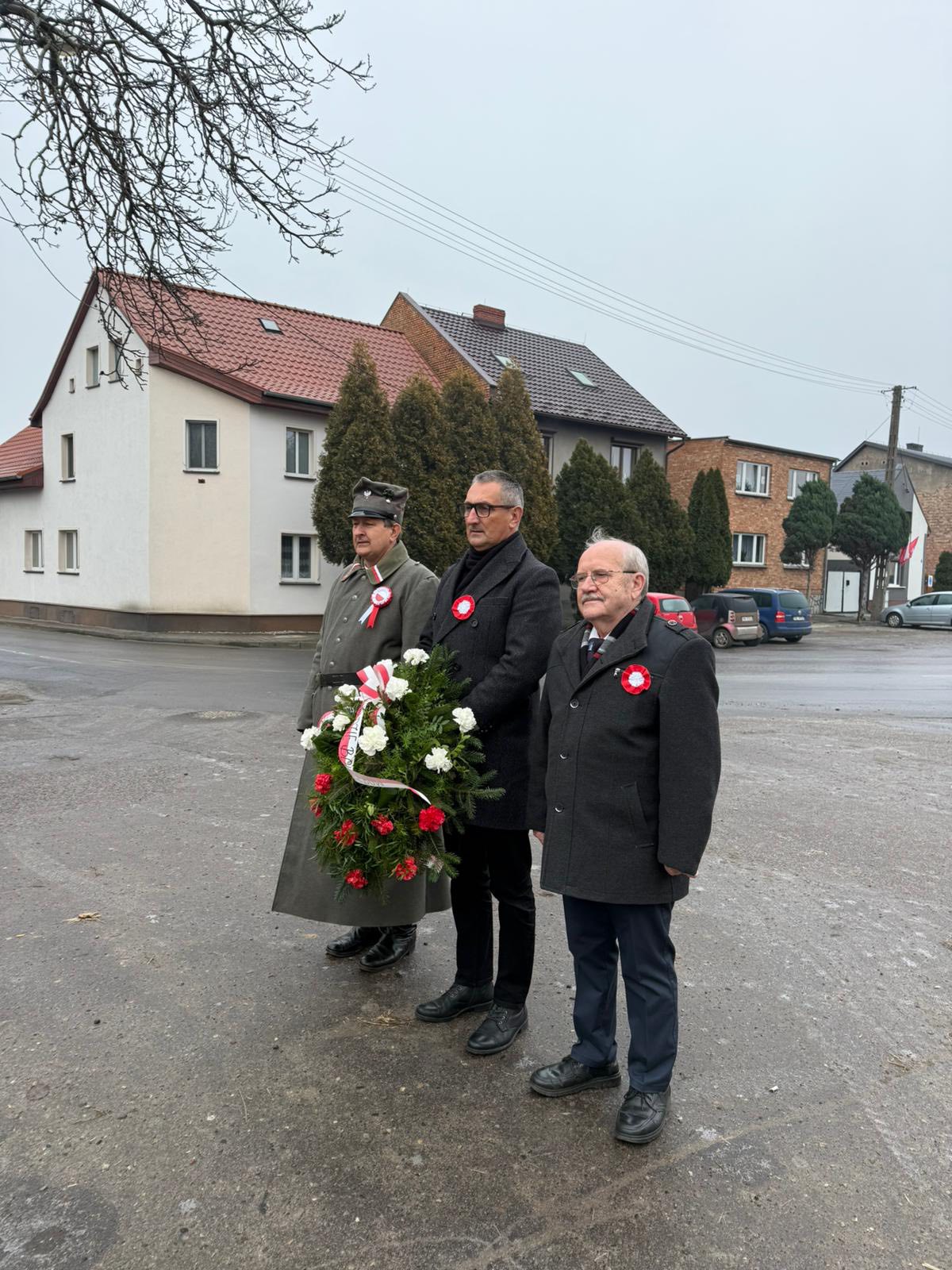 Na fotografii Janusz Frąckowiak, Wójt Gminy Przemęt, Robert Szwed, Prezes Towarzystwa Pamięci Powstania Wielkopolskiego – Koło w Przemęcie, oraz Antoni Fornalski, Honorowy Prezes Towarzystwa. Wójt Janusz Frąckowiak trzyma wiązankę, pan Robert i pan Antoni stoją obok. Wszyscy oddają hołd powstańcom przy pomniku na przemęckim rynku.