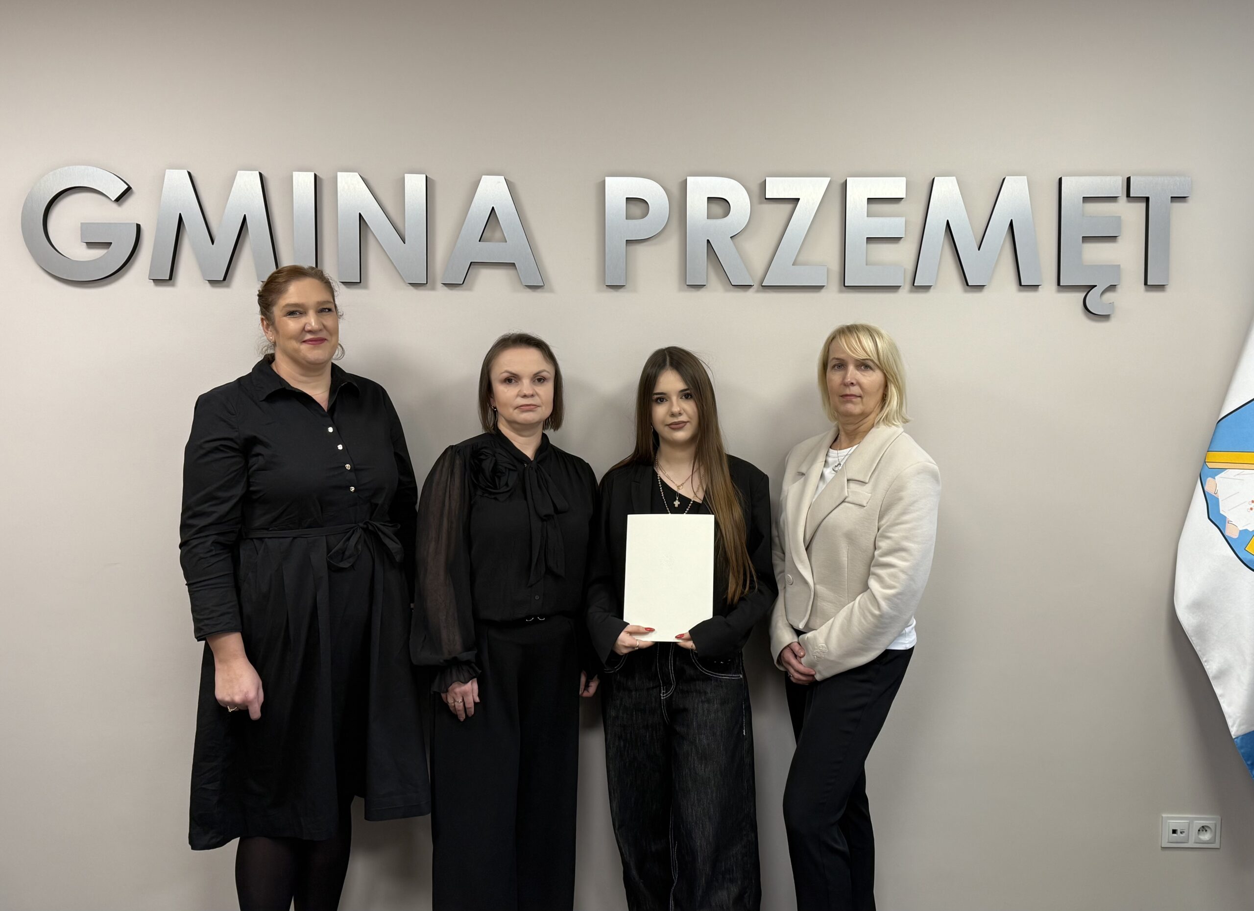 Na fotografii stypendystka wraz z mamą oraz pani kierownik wydziału oświaty i pani wicedyrektor szkoły ponadpodstawowej. Patrycja pozuje z dyplomem. W tle srebrny napis "Gmina Przemęt".
