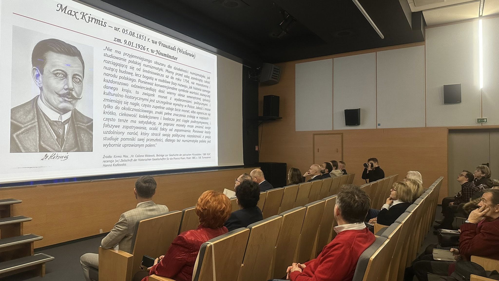 Na fotografii widoczne osoby siedzące na widowni i oglądające biografię Maxa Kirmisa, która wyświetlana jest na projektorze.